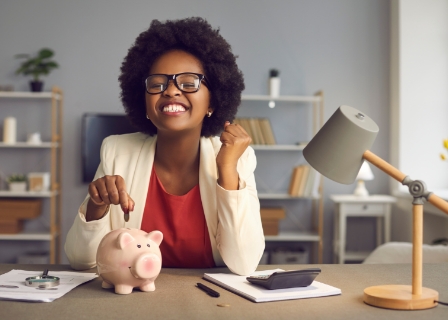a black woman with glasses in a home office smiles widely as she puts a coin in a piggy bank